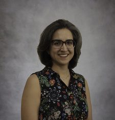 A woman with glasses standing in front of a gray background.